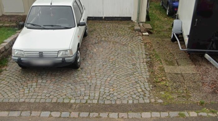 Uppfart med stensättning där en vit bil är parkerad bredvid ett hus och en släpvagn.