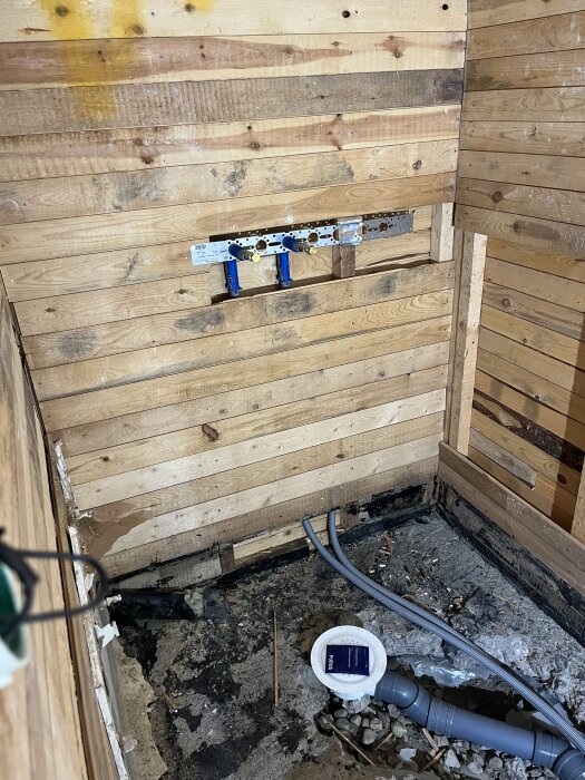 Grovt badrum under renovering med träpanel på väggarna och synliga rör och avlopp på det råa betonggolvet.