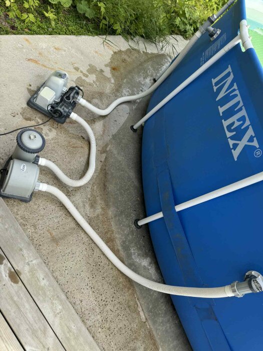 Utomhuspool från Intex med anslutna slangar och pumpar på en betongyta, omgiven av grönt lövverk.