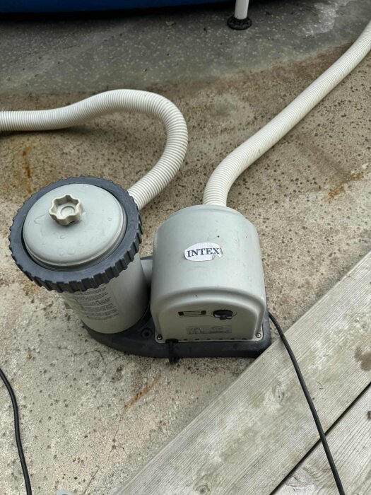 Grå Intex pump med filter och kopplade slangar på betongplatta, vid en träyta, relaterad till problem med installation och felmeddelande.