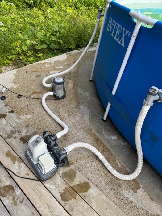 Uppblåsbar pool med två anslutna pumpar och slangar, stående på en altan med grön växtlighet i bakgrunden.