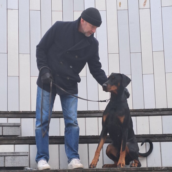 Man i mörk jacka klappar en svart och brun hund med koppel som sitter på Odenplan i Stockholm.