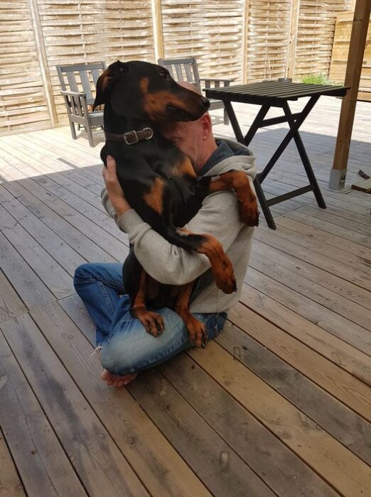 En man sitter på ett trädäck och kramar en stor hund som ligger i hans knä.