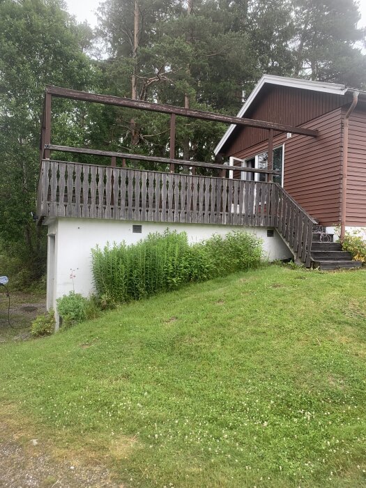 Trähus med en stor altan ovanpå en grässlänt, omgiven av träd och buskar i bakgrunden.