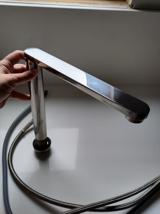 En modern köksblandare i rostfritt stål, hålls av en hand, visas tillsammans med anslutningsslangar på ett vitt underlag.