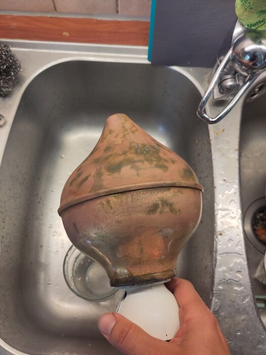 Knopp av förgylld koppar eller mässing hålls över en vask, visar patina och tecken på åldrande.