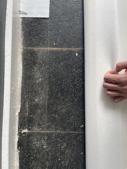 En hand håller i en vit skumrulle över ett svart golv med synlig tjärlim i skarvarna nära en list.