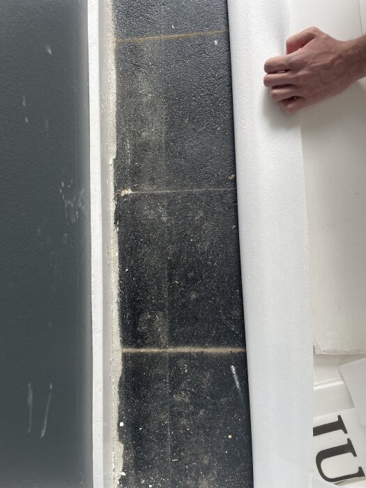 Golvyta med mörka plattor, delvis täckt av skummaterial, bredvid ett väggparti. En hand håller i skummaterialet.