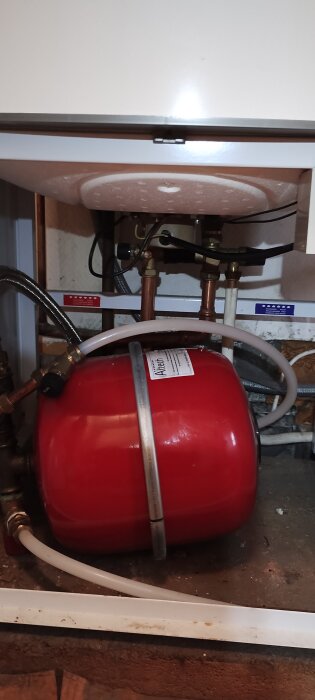 Röd expansionskärl under vit värmepanna med rör och slangar i ett installationsutrymme.