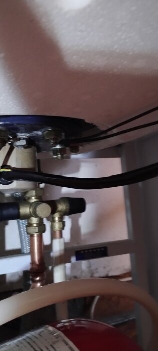 Närbild på rörkopplingar och ventiler under en panna, med kablar och en del av ett rött expansionskärl synligt nedtill.