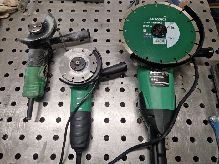 Tre vinkelslipar, två gröna med diamantklingor och en utan, ligger på ett metallbord med hål.