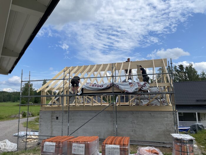 Bygget av ett takbjälklag med personer som snickrar på en ställning. Klar himmel i bakgrunden och omgivande natur.