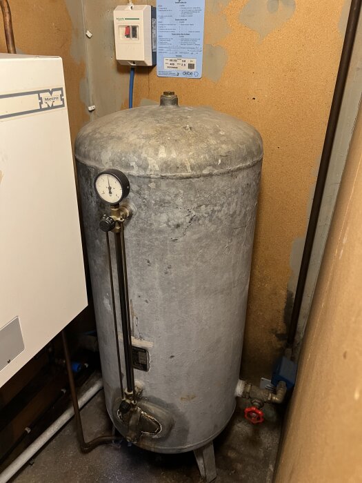 Grå trycktank med tryckmätare på sidan, rör och ventiler anslutna, samt en blå kontrollpanel på väggen bredvid.