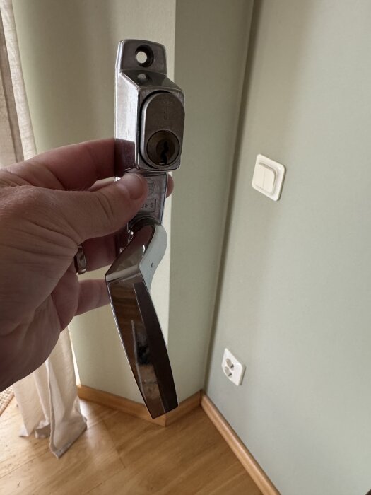 En hand håller ett Assa dörrhandtag med en låscylinder och två skruvhål för montering. Handtaget är borttaget från en glasdörr med aluminiumram.