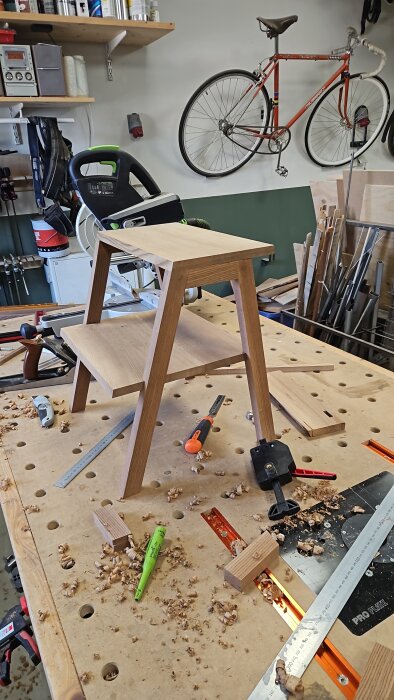 Träpall i alm på arbetsbänk i verkstad, omgiven av verktyg som såg, hyvel, och markeringspenna. Cykel och hyllor i bakgrunden.