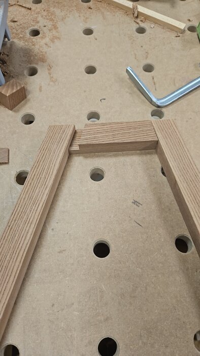 Träbitar av alm sammanfogade i hörnfogar på en arbetsbänk med hål, ett L-format verktyg syns bredvid.