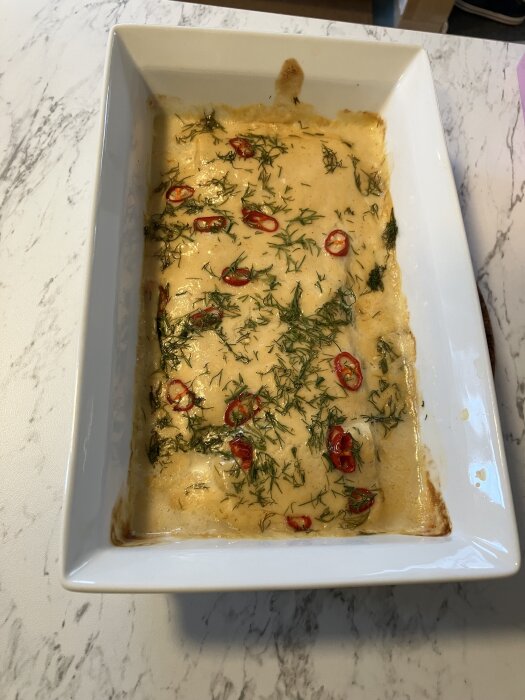 Ugnsform med torskrygg i lime- och chilisås, toppad med dill och skivad röd peppar på marmorbord.