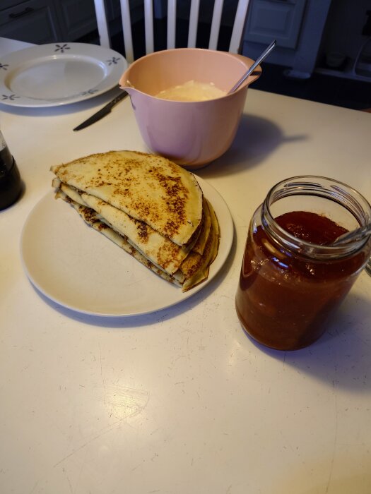 Pannkakor på en tallrik, en rosa skål med vispad grädde och en burk med sylt på ett bord i ett kök.
