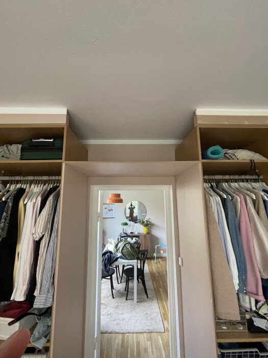 Inbyggda garderober längs en hallvägg med kläder hängt på klädstänger. Dörren leder vidare till ett matsalsrum med en spegel och bord.