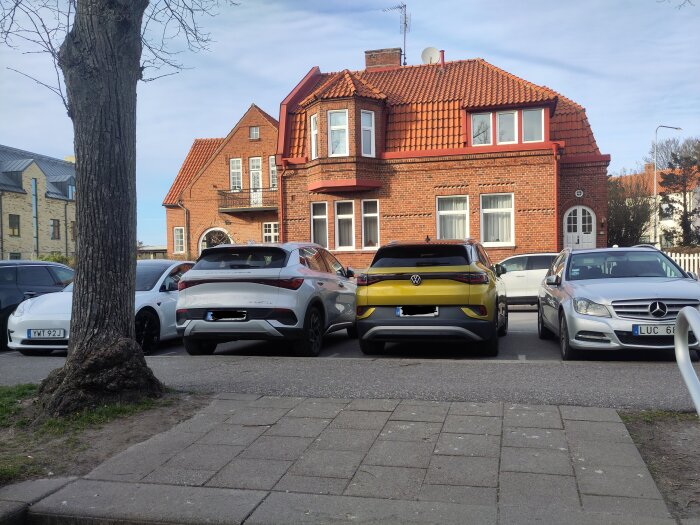 Flera bilar parkerade framför ett rött tegelhus, varav en är en gul Volkswagen SUV, med ett träd i förgrunden.