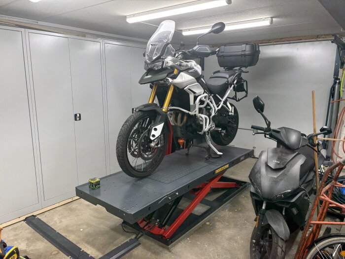 En motorcykel står på en höj- och sänkbar lyft i ett garage, redo för oljebyte och kedjeservice. En skoter syns också till höger.