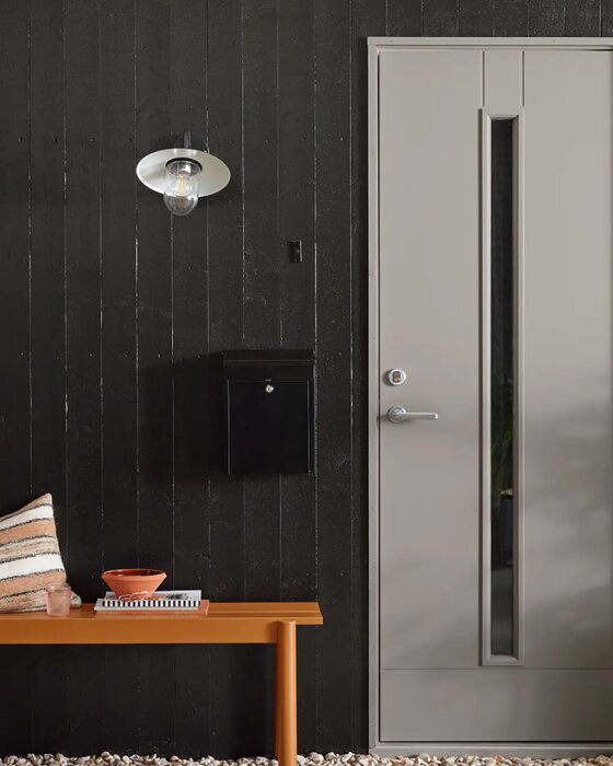 Svart träfasad med en beige dörr, modern utomhuslampa och svart brevlåda. En orange bänk med en kudde och skål står framför väggen.