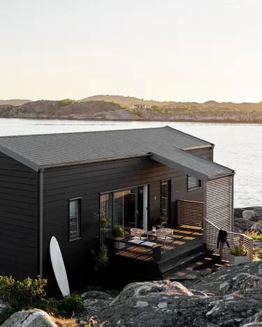 Svartmålad enplansstuga vid vattnet, med vit surfbräda lutad mot husväggen, träterrass och havsutsikt i bakgrunden.