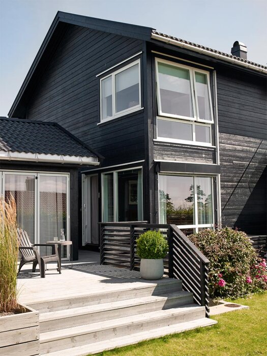 Svartmålat hus med vita knutar, trätrappa och uteplats med stolar. Grönska och rosor i trädgården.