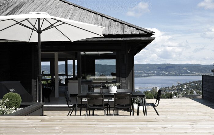 En fjällstuga med svartmålade väggar och vita knutar, stor uteplats med matbord, stolar och ett parasoll, utsikt över vattnet och skogsklädda kullar.