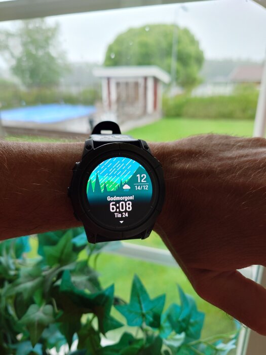 En smartklocka på en handled visar regnigt väder och tiden 6:08 medan handen hålls upp mot ett fönster med utsikt mot en trädgård.