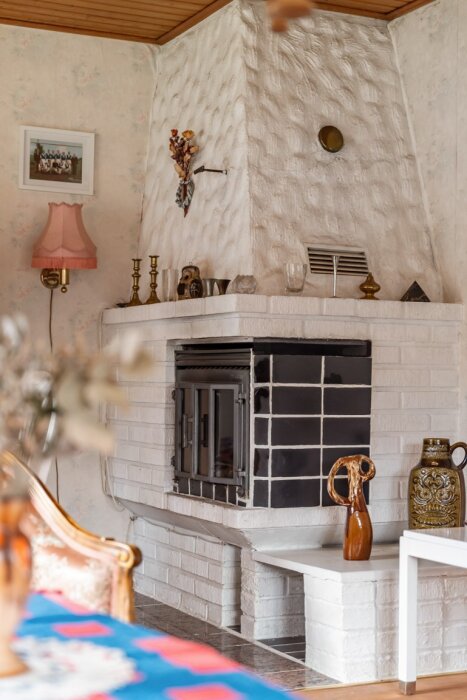 Öppen spis med vit murad yta och svart platt införing, dekorerad med vaser och ljusstakar i ett rum med blommigt tapet.