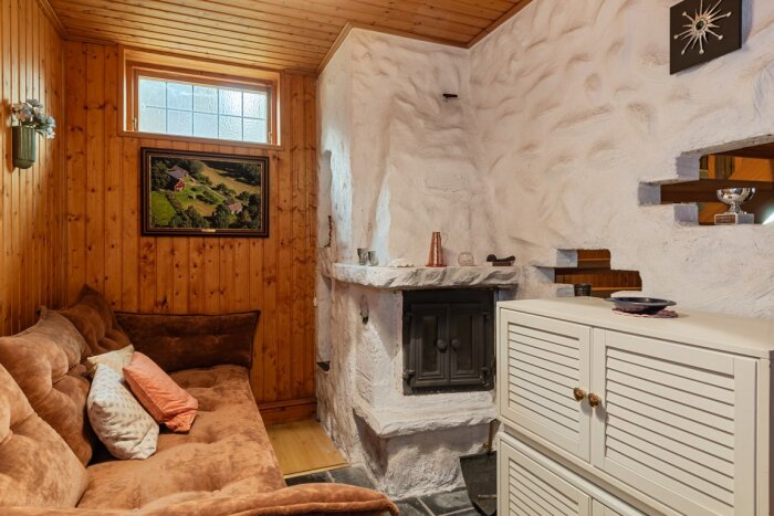 En rustik källare med en murad öppen spis, vitmålad, intill en brun soffa och ett skåp, med träpanel på väggarna och litet fönster ovan.