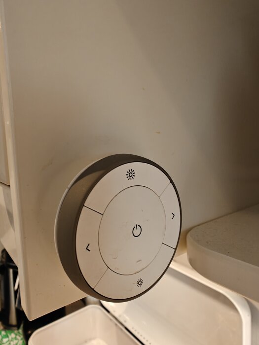 Vit rund IKEA-knapp för diskbänksbelysning på ett beige skåp, med olika symboler för ljusinställning.