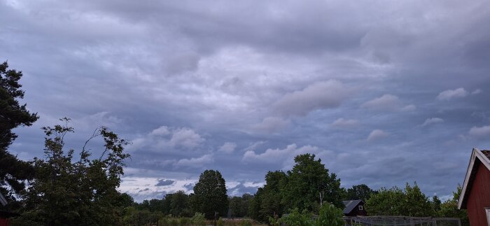 Molnig himmel över en lantlig trädgård med träd och en del av ett rött hus till höger.