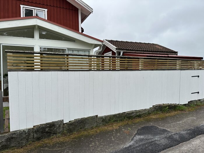 Vitt staket med nya ribbor framför ett rött hus. Staketet har en stenmur som grund och ligger bredvid en asfalterad väg.