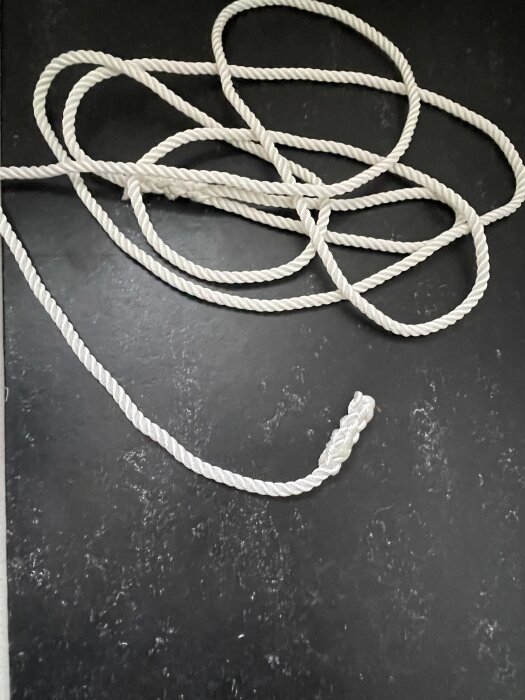 En vit repända ligger böjd och spiralformad på en svart yta. Repet har en knut i ena änden.