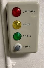 Kontrollpanel med röd, gul och grön lampa, samt en tryckknapp, markerad med orden "Upptagen", "Vänta", "Stig in", och "Tryck".