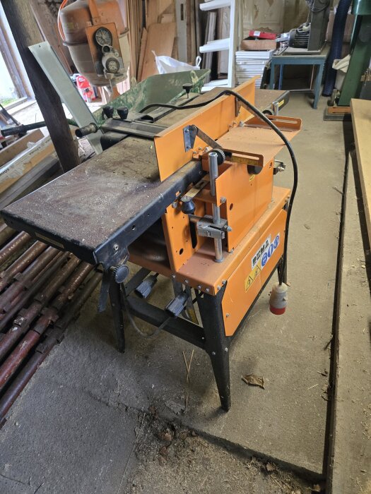 Orange träbearbetningsmaskin i verkstadsmiljö med verktyg och material i bakgrunden.