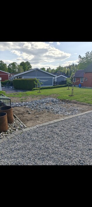 Gräsyta med jord och grus förberett för stensättning, omgiven av hus och trädgårdar på en solig dag.