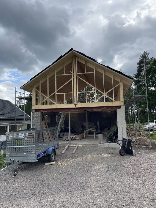 Trästruktur av ett pågående byggprojekt med takpapp; släp och byggmaterial i förgrunden, molnig himmel i bakgrunden.