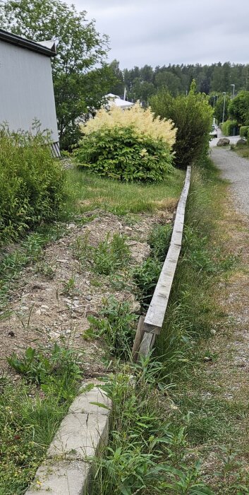 Trädgård med ett lutande träräcke som tryckts ut av jord, med en grävd ränna längs räcket och en stenmurskant synlig bredvid.