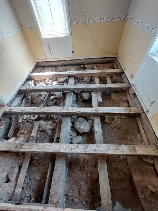 Renoveringsrum med exponerade golvbjälkar och brädor ovan en jordgolv. Murstocken syns i bakgrunden. Ljust fönster till vänster.