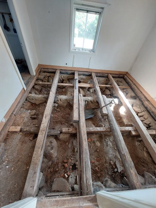 Rivna golvbrädor i ett renoveringsprojekt, visar bjälklag och stenar i torpargrund, med ett fönster i bakgrunden och en spade på marken.