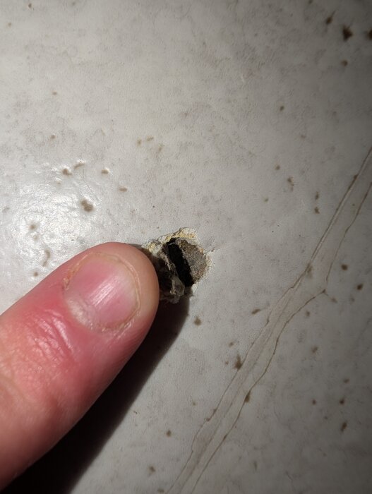 Hål i en ljus plastmatta i köket, med en finger som pekar på vad som ser ut som mörkt material under, eventuellt svartlim.