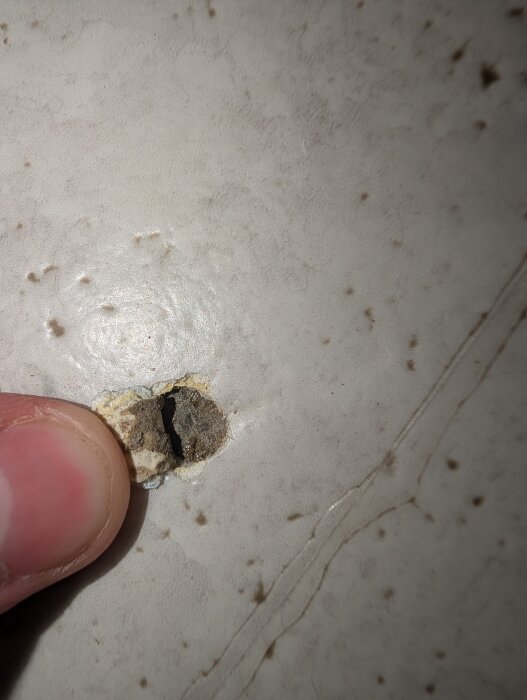 Hål i gammal plastmatta i köket, finger pekar på misstänkt svartlim synligt under ytan.