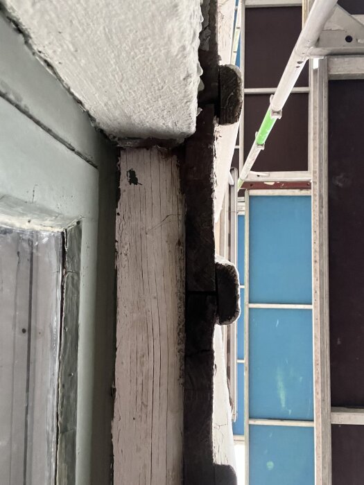 Källarfönster sett underifrån där träpanelen möter en putsad mur, med byggnadsställning i bakgrunden.