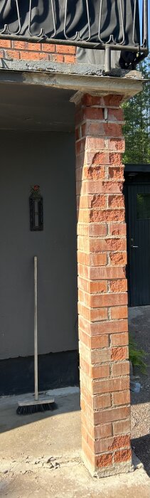 Tegelstolpe vid husfasad med slitage och sprickor, kvast står lutad mot väggen.