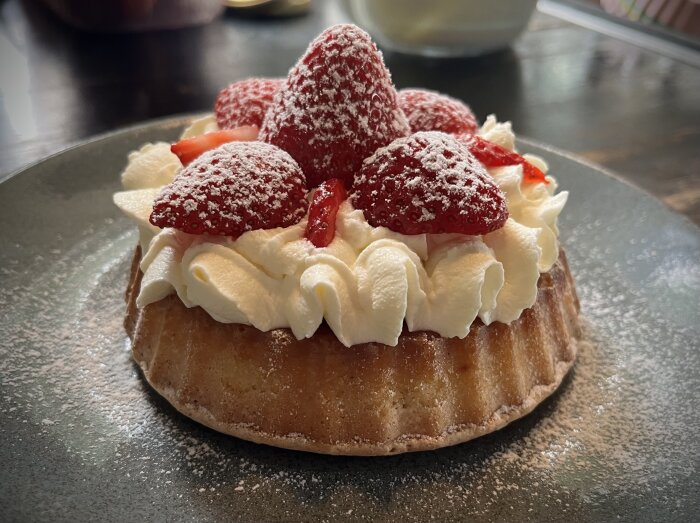 Bakelse med vispad grädde och jordgubbar på en tartlettform, lätt pudrad med florsocker, serverad på en tallrik.