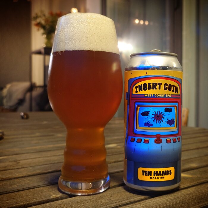 Ett glas fyllt med bärnstensfärgad öl bredvid en ölburk märkt "Insert Coin" från Ten Hands Brewing på ett träbord.