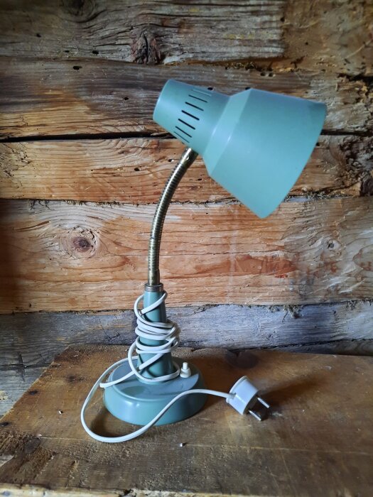 Grön bordslampa av äldre modell med flexibel arm och ojordad kontakt, placerad mot en trävägg.